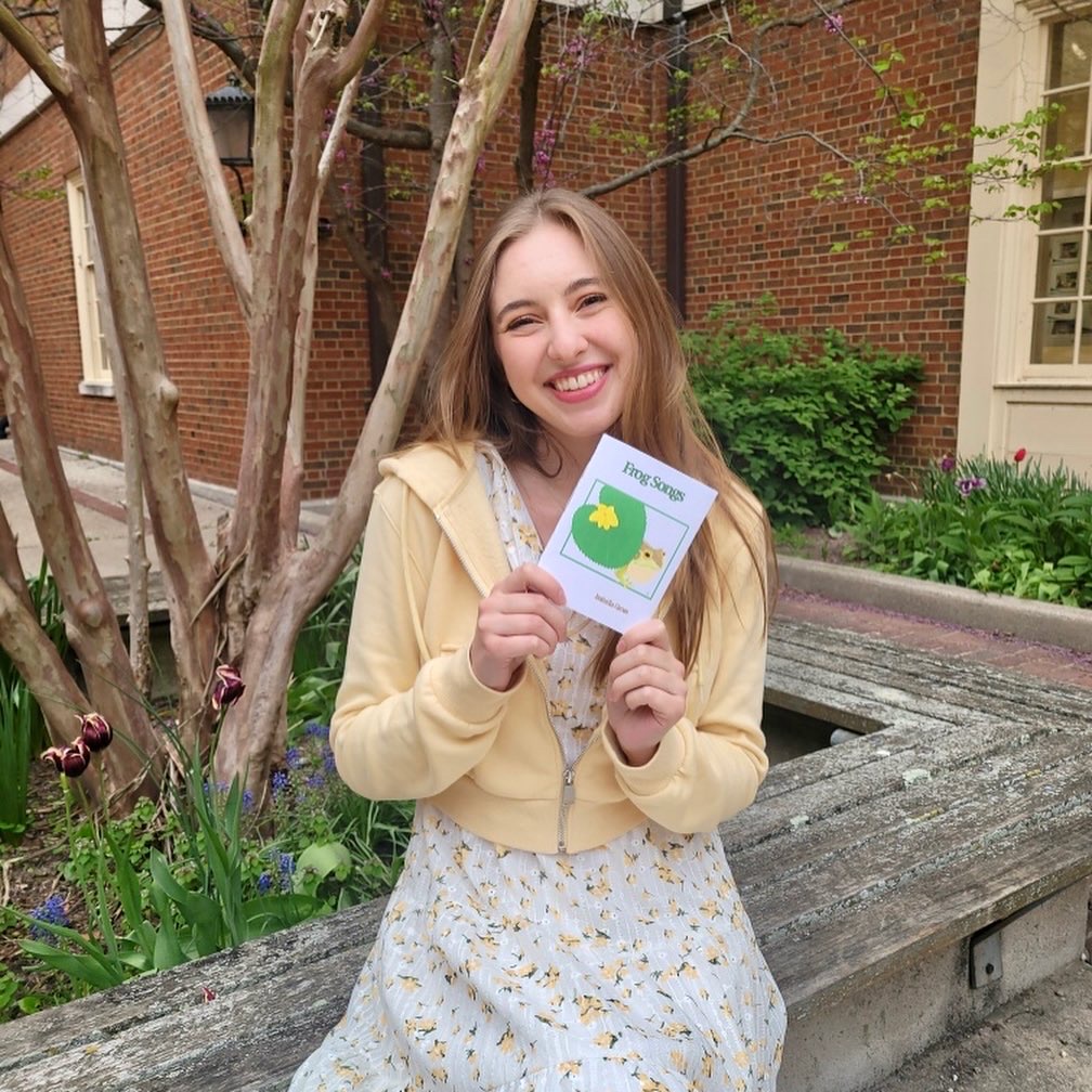 Isabella Gross with her first handmade book, titled Frog Songs, seated in a courtyard in spring.